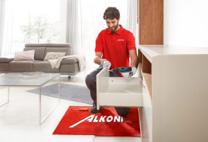 Mann in rotem Hemd beim Zusammenbau von Möbeln in einem hellen Wohnzimmer, kniend auf einer roten ALKONI-Matte, Werkzeuge in der Nähe.